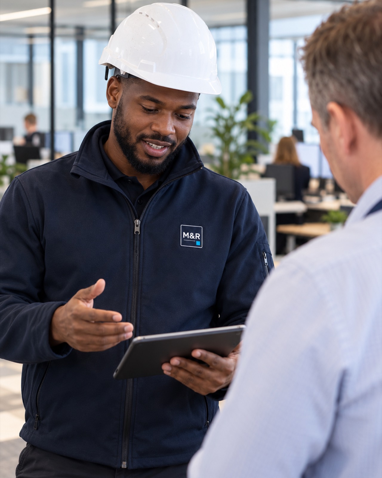 Kwame Asante, project manager, in a hard hat and a Marlow & Reed jacket, talking a client through a tablet in an open-plan office.