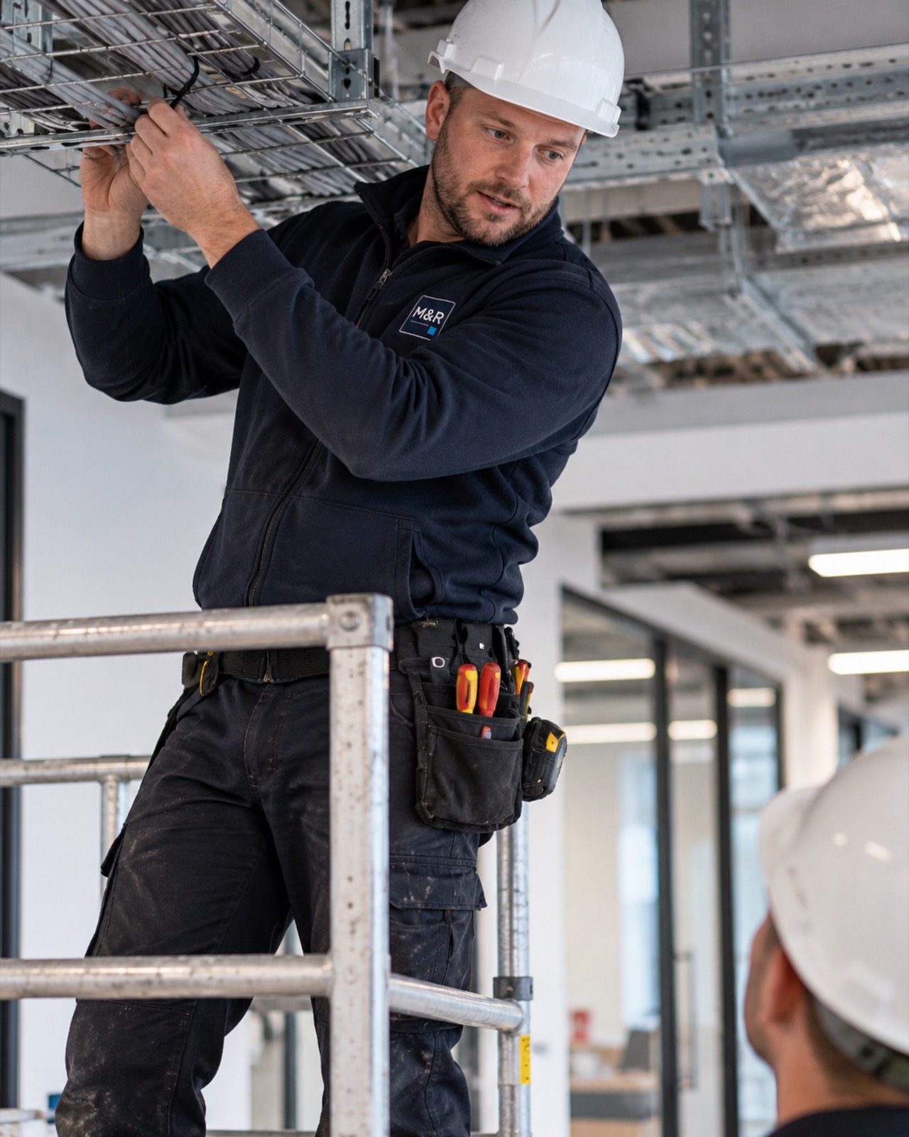 Liam Brady, electrical supervisor, on a podium step fixing cable tray in an office ceiling, wearing a hard hat and a tool belt.
