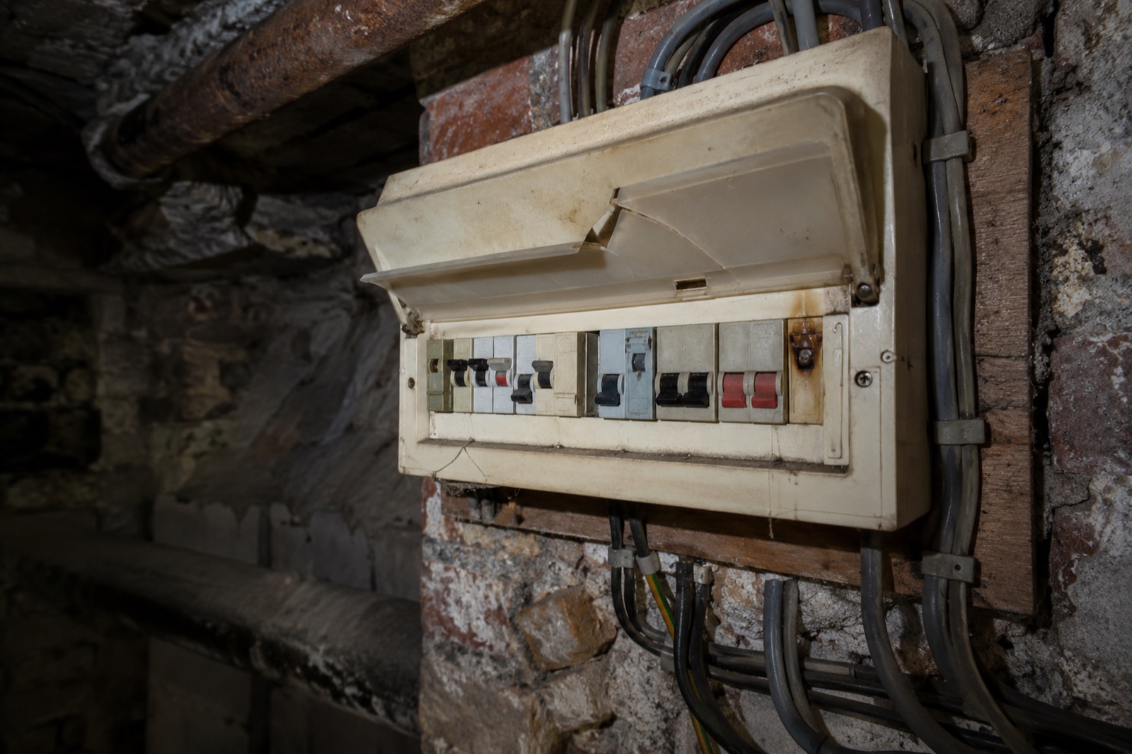 An old cream consumer unit in a brick cellar, its cover cracked, with mismatched breakers and a scorch mark by one terminal.