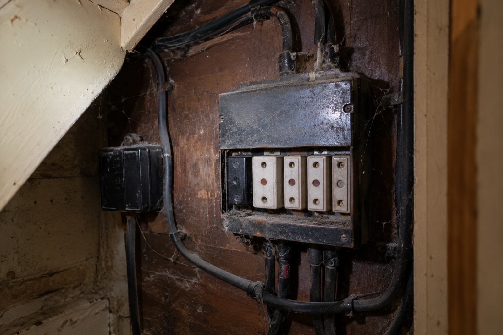 An old fuse box with rewireable fuses on a dark wooden board under the stairs, black rubber cables and cobwebs.