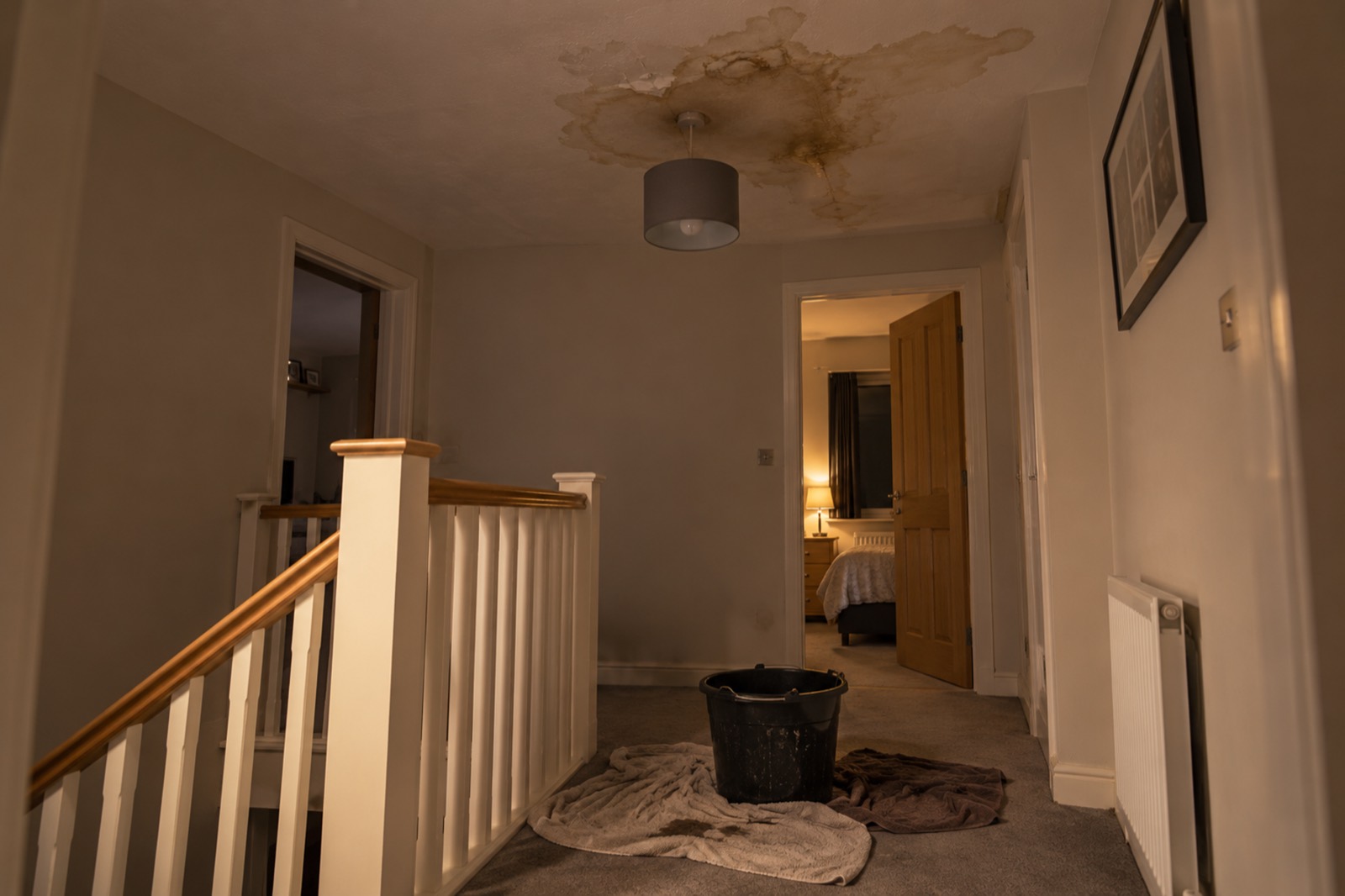 A landing at night, its ceiling stained around the light fitting, with a bucket and towels on the carpet below.