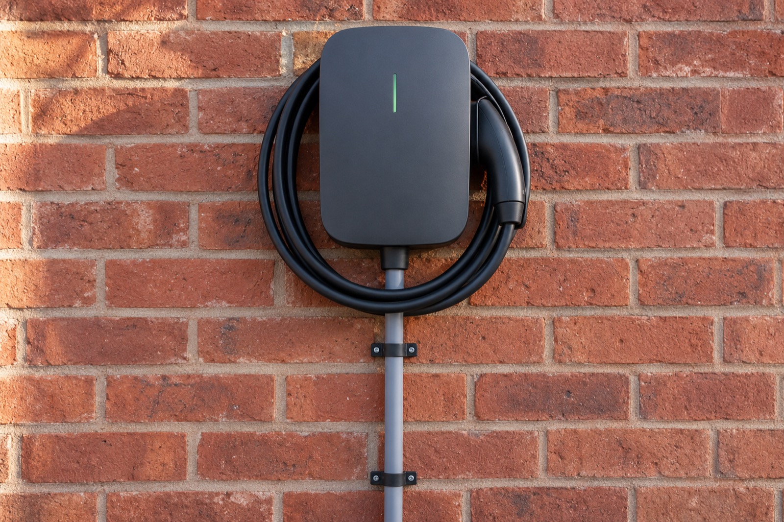 A charcoal wall charger on red brick with its black cable coiled round it and a grey conduit entering from below.