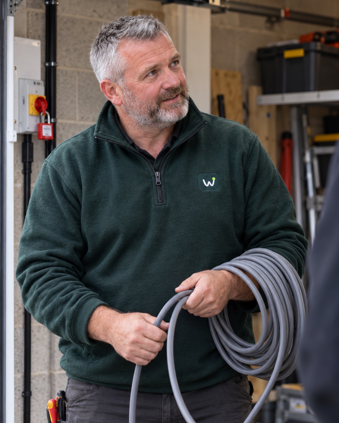Matt in a garage with a coil of grey cable over his arm, looking up to talk, an isolator with a red lock-off behind him.